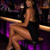 A woman poses playfully at a bar, wearing a black outfit and high heels, with colorful lighting in the background.
