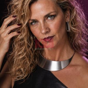 A woman with curly hair and bold makeup poses elegantly, wearing stylish jewelry and a sleek black outfit.