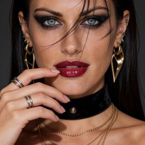 Ritratto di una donna con trucco intenso, labbra rosse, collana nera e orecchini dorati. Capelli scuri e lisci.