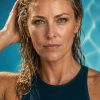 A close-up portrait of a woman with wet hair, wearing a dark swimsuit, against a blue background.