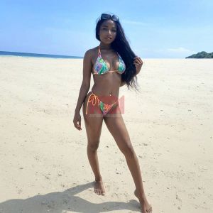 A person in a colorful bikini stands on a sandy beach under a clear blue sky. Sunlight reflects off the ocean.