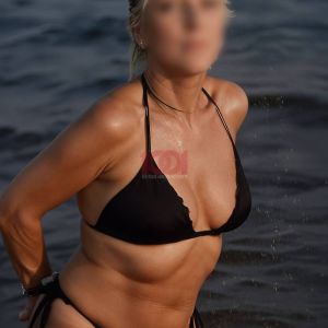 Una donna bionda indossa un bikini nero mentre si trova vicino all'acqua. Sole e onde sullo sfondo.