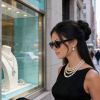Donna elegante in abito nero e occhiali da sole, osserva gioielli in vetrina di Tiffany & Co. con caffè e cornetto.