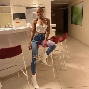 A stylish woman poses in a kitchen, wearing a white top, ripped jeans, and white boots, with a drink nearby.