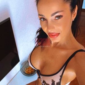 A woman poses for a selfie, wearing lingerie with decorative elements in a cozy indoor setting.