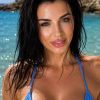 A woman with wet hair in a blue bikini poses confidently on a beach with clear blue water in the background.