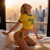 A woman poses in a yellow Brazil shirt and bikini, illuminated by a sunset with a city skyline in the background.