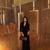 A woman in a black dress poses elegantly in front of a shimmering golden curtain. She holds a stylish clutch.