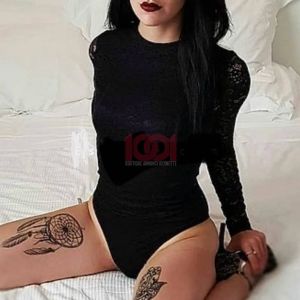 Giovane donna vestita di nero, con tatuaggi su cosce, seduta su un letto bianco, sguardo intenso e trucco scuro.