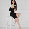 A stylish woman poses on a stool, wearing a black dress and silver high heels, holding flowers in a bright room.