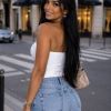 A woman smiles over her shoulder, wearing a white top and blue jeans, against a city street background.