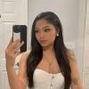 A woman takes a mirror selfie, wearing a white top, with long hair, and makeup enhancing her features.