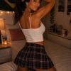 A person poses playfully on a bed, wearing a crop top and plaid skirt, with warm lighting and a cozy background.