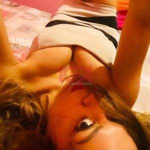 A person is lying on a bed with a pink background, wearing a fitted outfit and striking a playful pose.