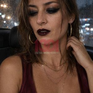 Donna con trucco scuro e labbra bordeaux, con vestito violaceo e gioielli, seduta in un'auto con finestra bagnata.