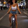 A woman rides a bike at night, smiling, wearing a crop top and shorts, with a vibrant street scene around her.