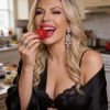 Donna sorridente in cucina, indossa un accappatoio nero e orecchini, tiene una fragola da mangiare.