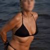 Una donna bionda indossa un bikini nero mentre si trova vicino all'acqua. Sole e onde sullo sfondo.