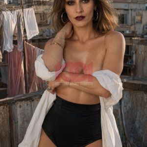 Donna con capelli ondulati, top bianco e pantaloni neri, in piedi su un tetto con edifici sullo sfondo.