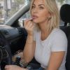 Una donna bionda si trucca le labbra con smalto in un'auto. Indossa una maglietta bianca e jeans.