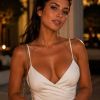 A woman in a white dress smiles softly, holding a stylish handbag, with warm, soft lighting in the background.