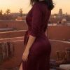 A woman in a burgundy dress stands on a rooftop at sunset, with a cityscape and palm trees in the background.