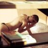 A woman with a shaved head poses on a bed, partially obscured by a table and surrounded by books.