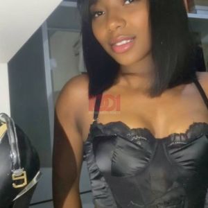 A young woman poses with a smile, wearing a black top, in a stylish room with various accessories visible.