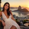 A woman in a white dress poses elegantly against a stunning sunset over a coastal city view.