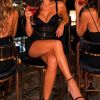 A glamorous woman sips a cocktail in a stylish bar, with city lights twinkling in the background at night.