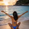 A person in a blue bikini sits on the beach, arms outstretched, enjoying a sunset over the water.