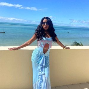 A woman in a light blue dress poses by the ocean under a clear blue sky, enjoying a sunny day.