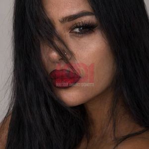 A close-up portrait of a woman with long black hair, striking makeup, and a focused expression against a neutral background.