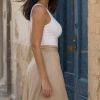 A woman with long hair in a white top and beige skirt stands beside a blue door in a sunlit street.