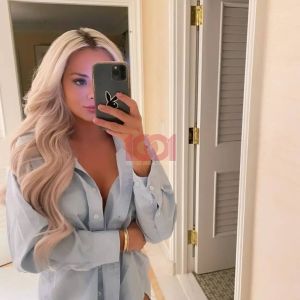 A woman with wavy blonde hair takes a mirror selfie wearing an unbuttoned blue shirt. Casual indoor setting.