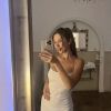 A person poses for a selfie in a white dress, reflecting in a mirror with elegant decor in the background.