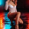 A woman poses in a stylish, fishnet bodysuit and heels, illuminated by colorful lights on a reflective floor.