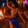 A vibrant beach party scene at night with dancing people, a bonfire, and a woman in a sparkling blue outfit.