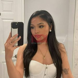 A woman takes a mirror selfie, wearing a white top, with long hair, and makeup enhancing her features.