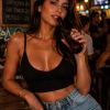 A woman with dark hair poses with a beer bottle in a lively bar setting, wearing a black top and denim shorts.