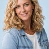 A smiling woman with curly hair wears a denim jacket over a white shirt, standing against a light blue background.