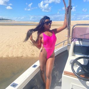 A woman in a bright pink swimsuit poses on a boat by a sandy beach under a clear blue sky.