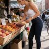 Donna con capelli ricci e pantaloni attillati, sceglie pesche in un mercato all'aperto. Scarpe bianche.
