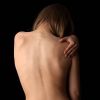 A back view of a person with long hair, gently holding their shoulder, against a dark background.