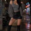 Una donna in outfit elegante, con giacca oversize e stivali alti, posa in un ambiente urbano illuminato.