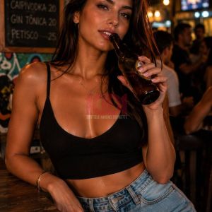 A woman with dark hair poses with a beer bottle in a lively bar setting, wearing a black top and denim shorts.