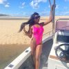 A woman in a bright pink swimsuit poses on a boat by a sandy beach under a clear blue sky.