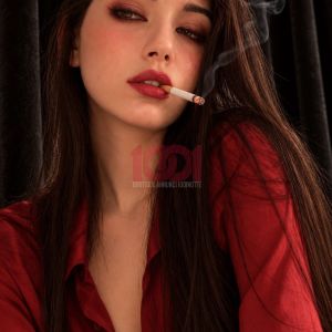 Donna dai capelli lunghi e scuri, con una camicia rossa e una sigaretta, posa elegantemente davanti a un fondale nero.