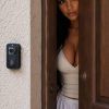 A person peeks through a partially open door, wearing a light top and shorts, with a curious expression.