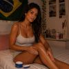 A woman applies lotion to her leg while sitting on a bed with soft lighting and a Brazilian flag in the background.
